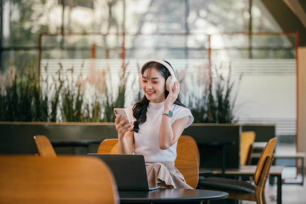 Young Asian freelancer woman is listening to music on her wireless headphones and working on her laptop in a coworking space, holding her phone in her hand and smiling